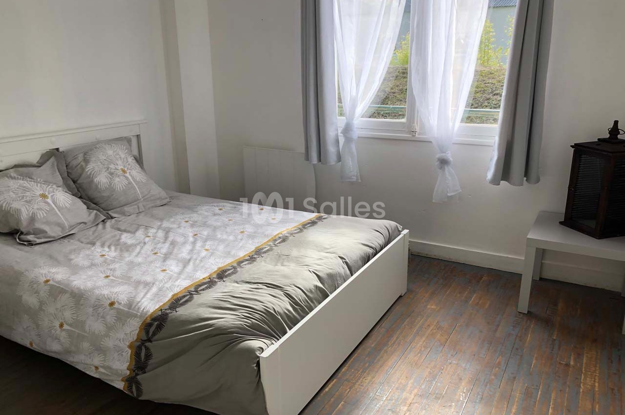 Chambre lumineuse avec lit double, draps à motifs, parquet en bois et fenêtre avec rideaux blancs.