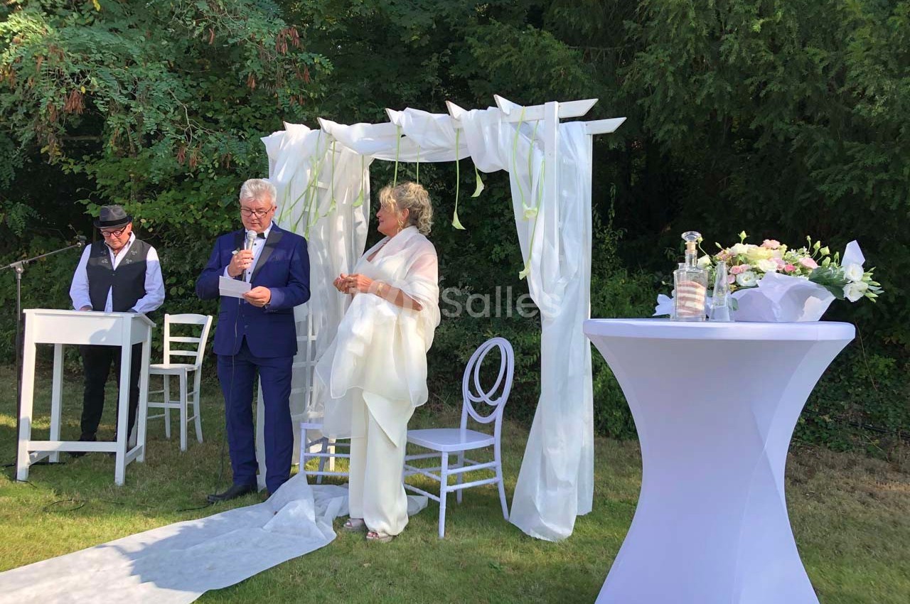 Un couple échange des vœux sous une arche blanche décorée, dans un jardin, avec un officiant à leurs côtés.