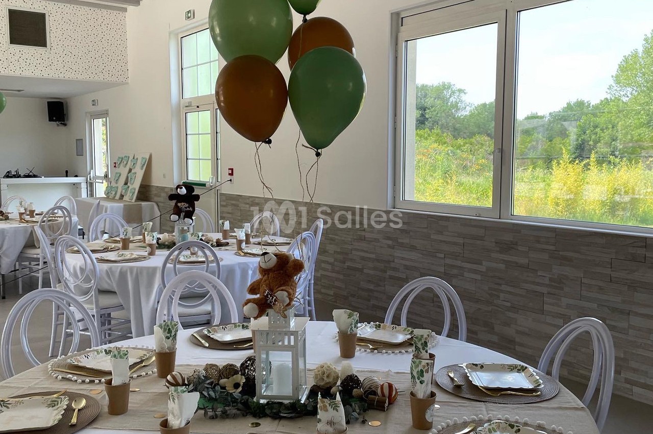 Salle décorée pour un événement avec des tables rondes dressées, des ballons verts et marron, et une vue sur l'extérieur…