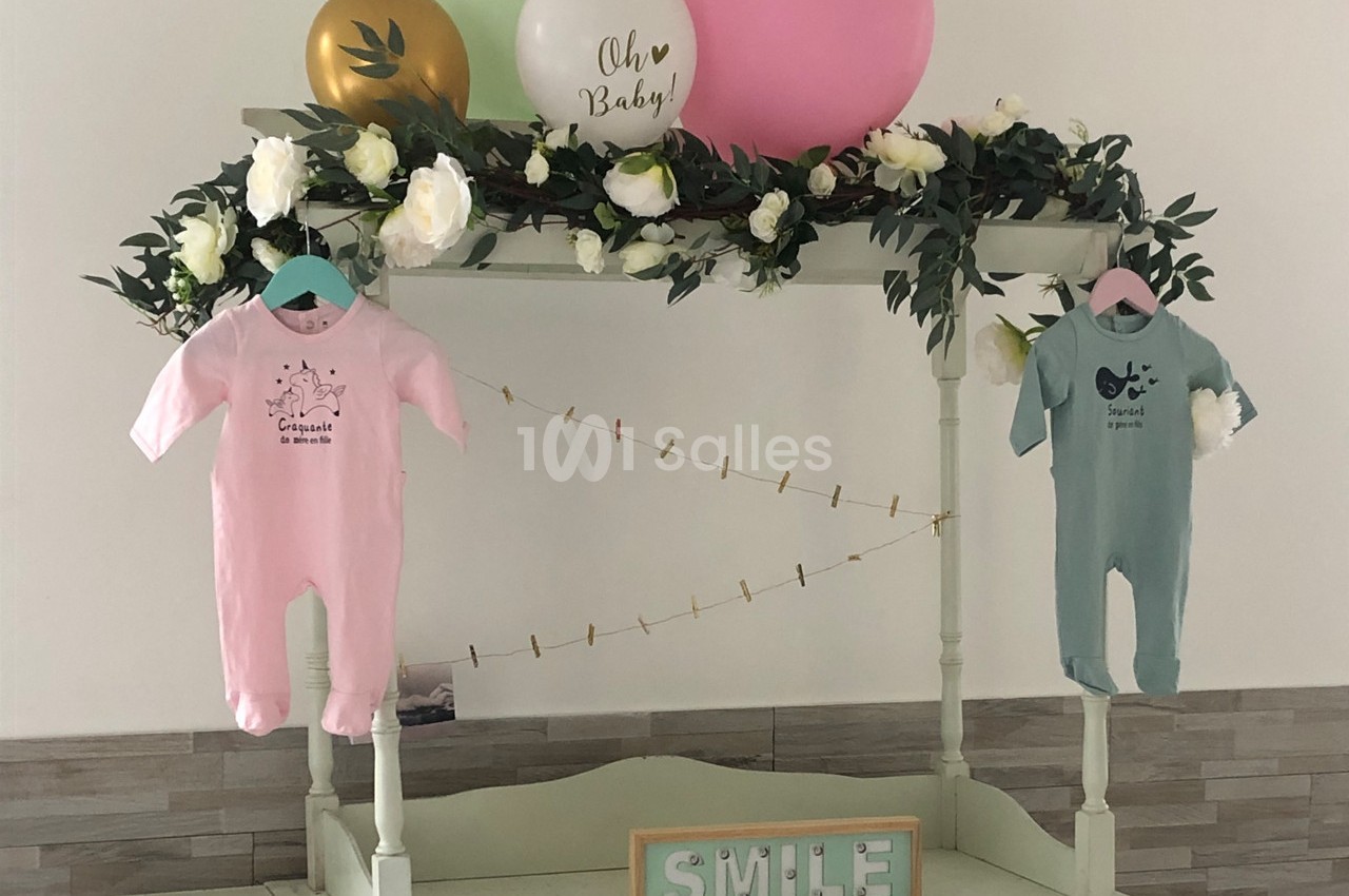 Deux grenouillères suspendues, rose et verte, décorées avec des fleurs, ballons et une pancarte ’Smile’.