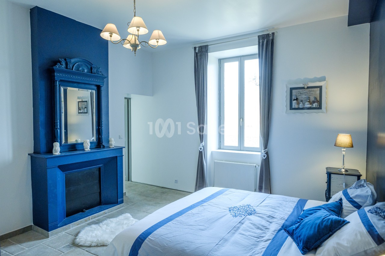 Chambre lumineuse avec murs blancs, cheminée bleue, lit double et grande fenêtre avec rideaux gris.