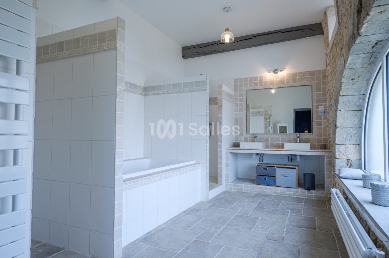 Salle de bain lumineuse avec baignoire, grand miroir, murs en carrelage beige et mur en pierre avec fenêtre cintrée.