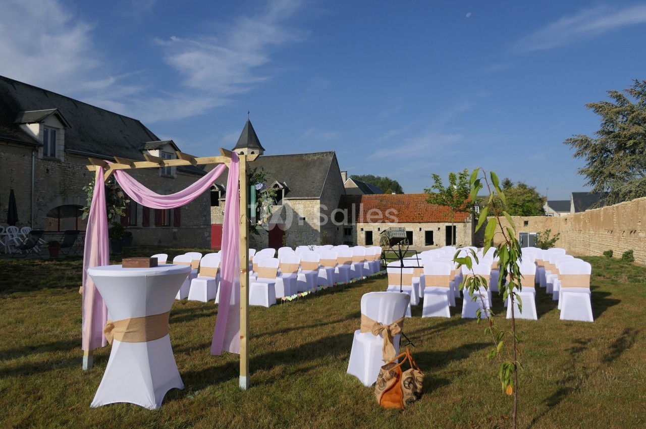 Chaises blanches alignées en extérieur pour une cérémonie, avec arche décorée et bâtiments en pierre en arrière-plan.