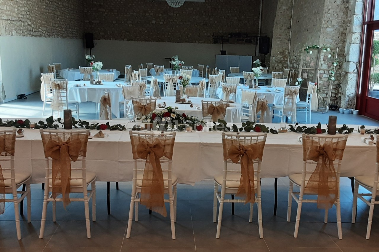 Salle de réception décorée pour un événement, avec tables dressées, chaises ornées de nœuds en toile de jute et compositions…