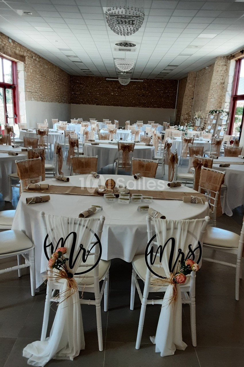 Salle de réception décorée pour un mariage, avec tables dressées, chaises ornées et lumière naturelle.