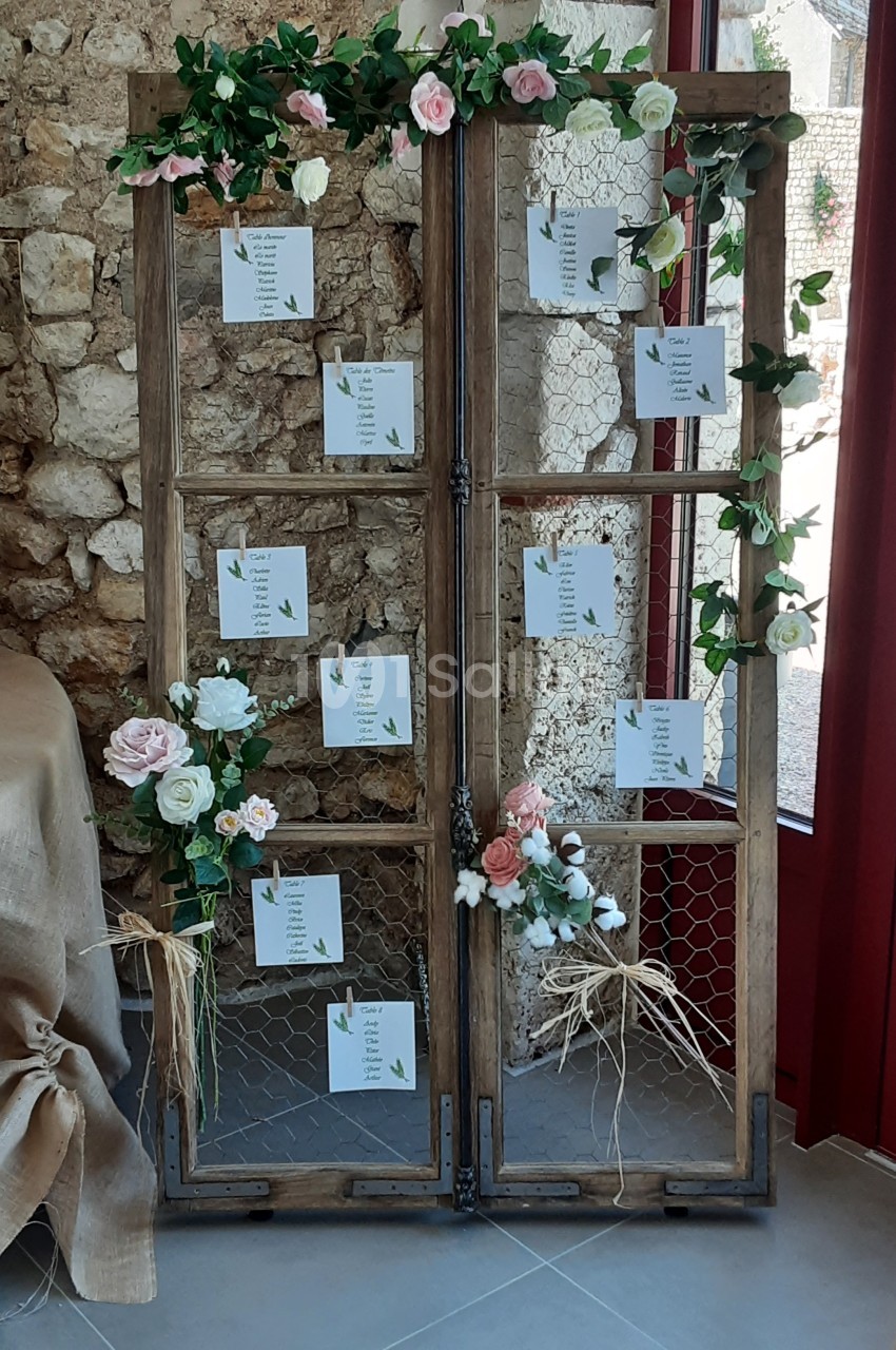 Un plan de table de mariage sur une structure en bois décorée de fleurs et de feuillages, placé près d'un mur en pierre.