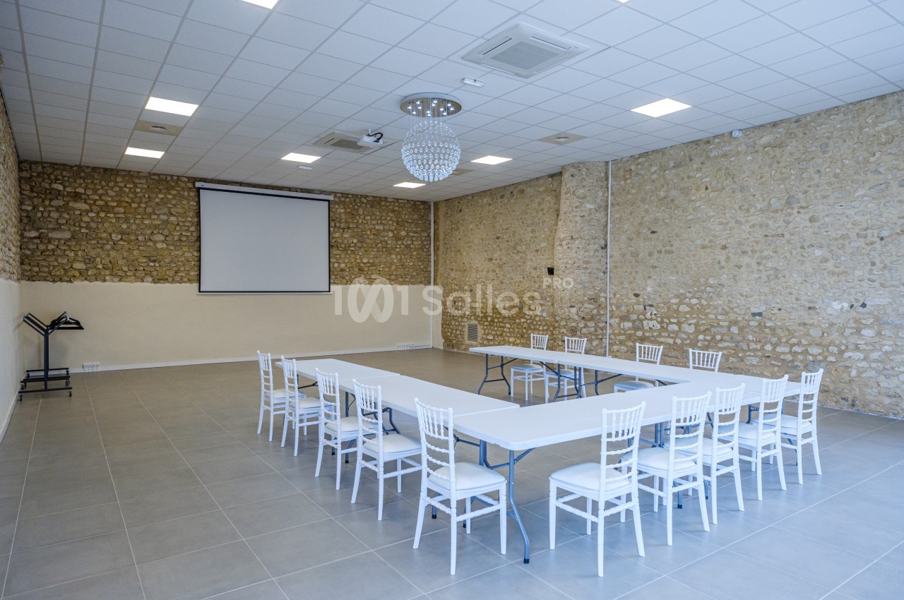 Salle lumineuse avec murs en pierre, tables blanches disposées en U, chaises blanches et écran de projection.