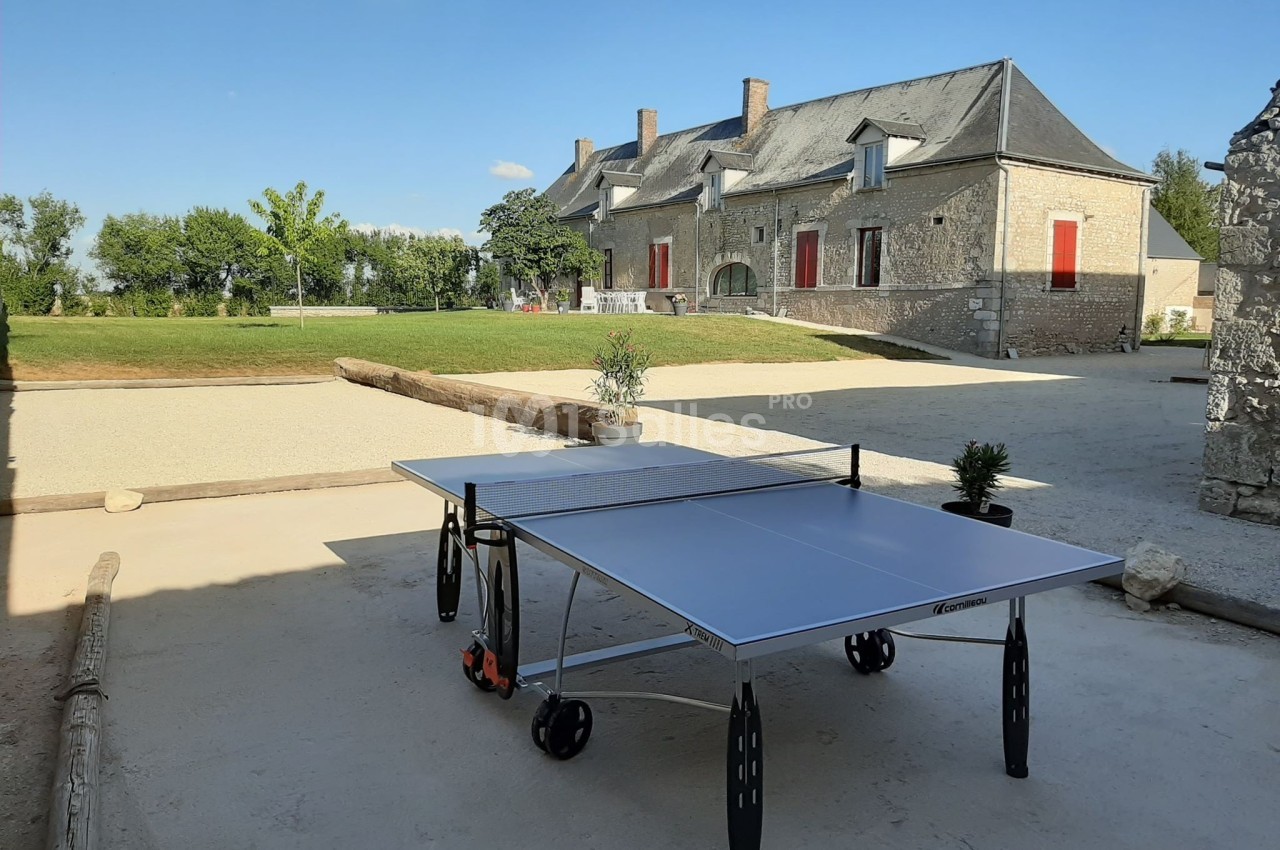 Table de ping-pong installée dans une cour ensoleillée avec une bâtisse en pierre et un jardin en arrière-plan.
