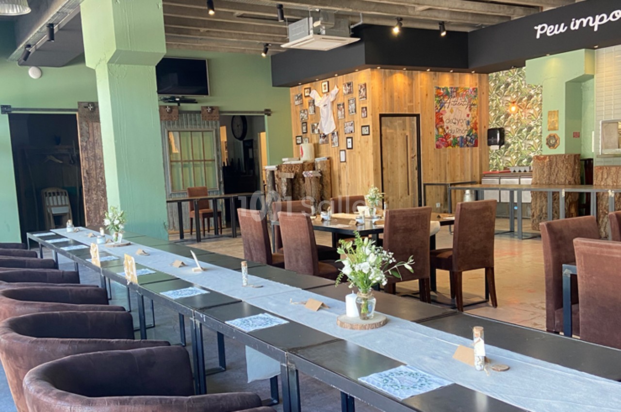 Salle de restaurant lumineuse avec de longues tables dressées, des chaises en velours et une décoration murale éclectique.