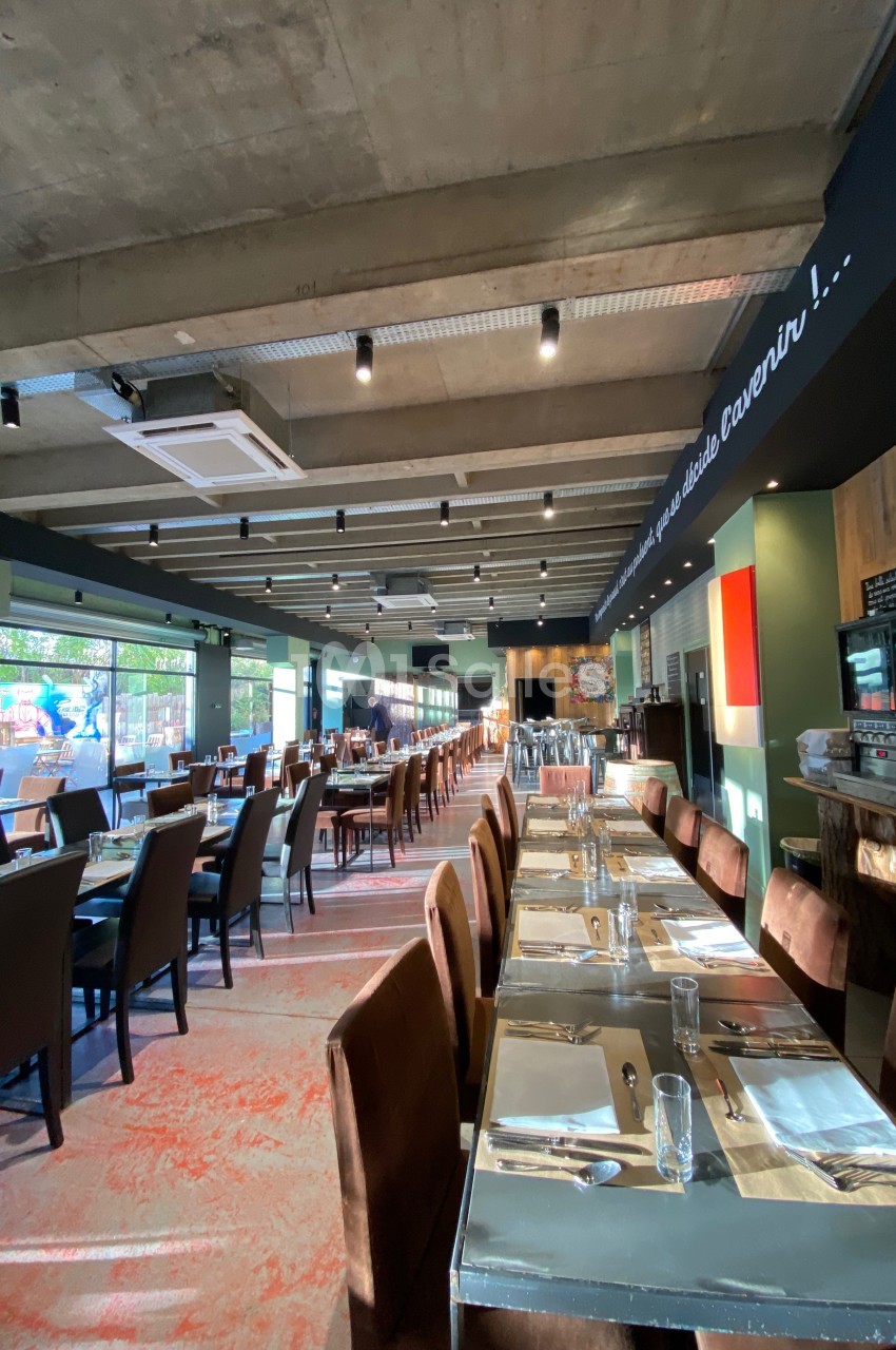 Salle de restaurant lumineuse avec tables dressées, grandes baies vitrées et plafond en béton apparent.