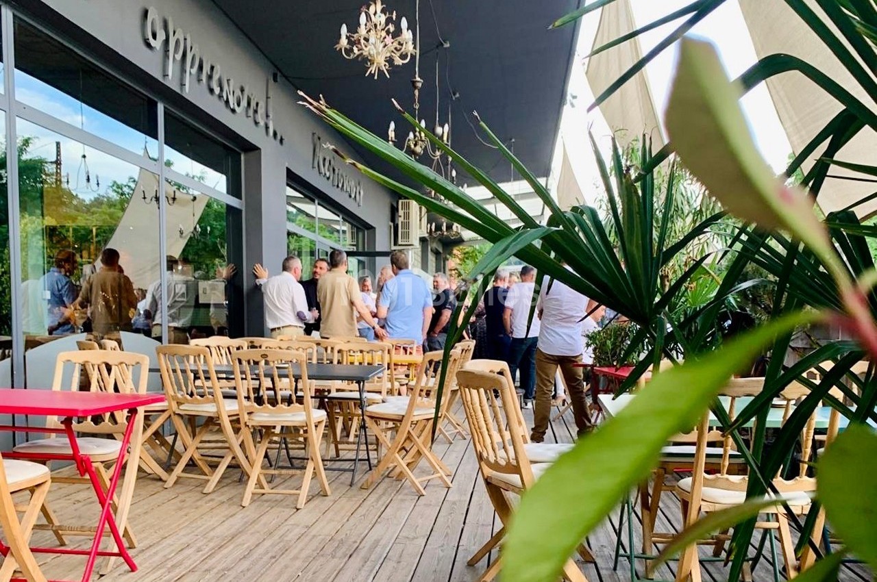 Terrasse en bois avec des tables et chaises, des plantes en premier plan, et un groupe de personnes discutant au fond.