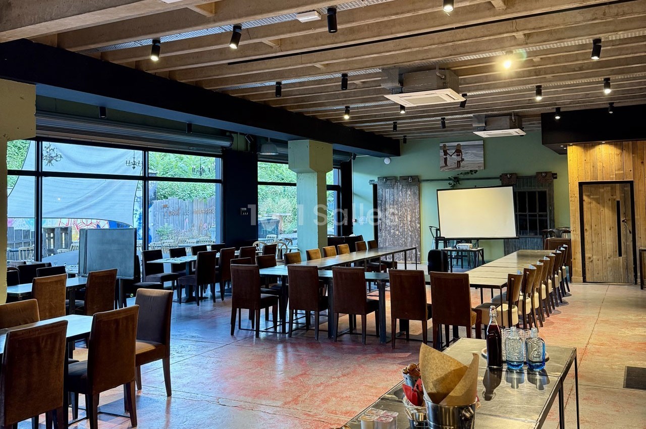 Salle de restaurant spacieuse avec de longues tables en bois, chaises alignées, et un grand écran blanc au fond.