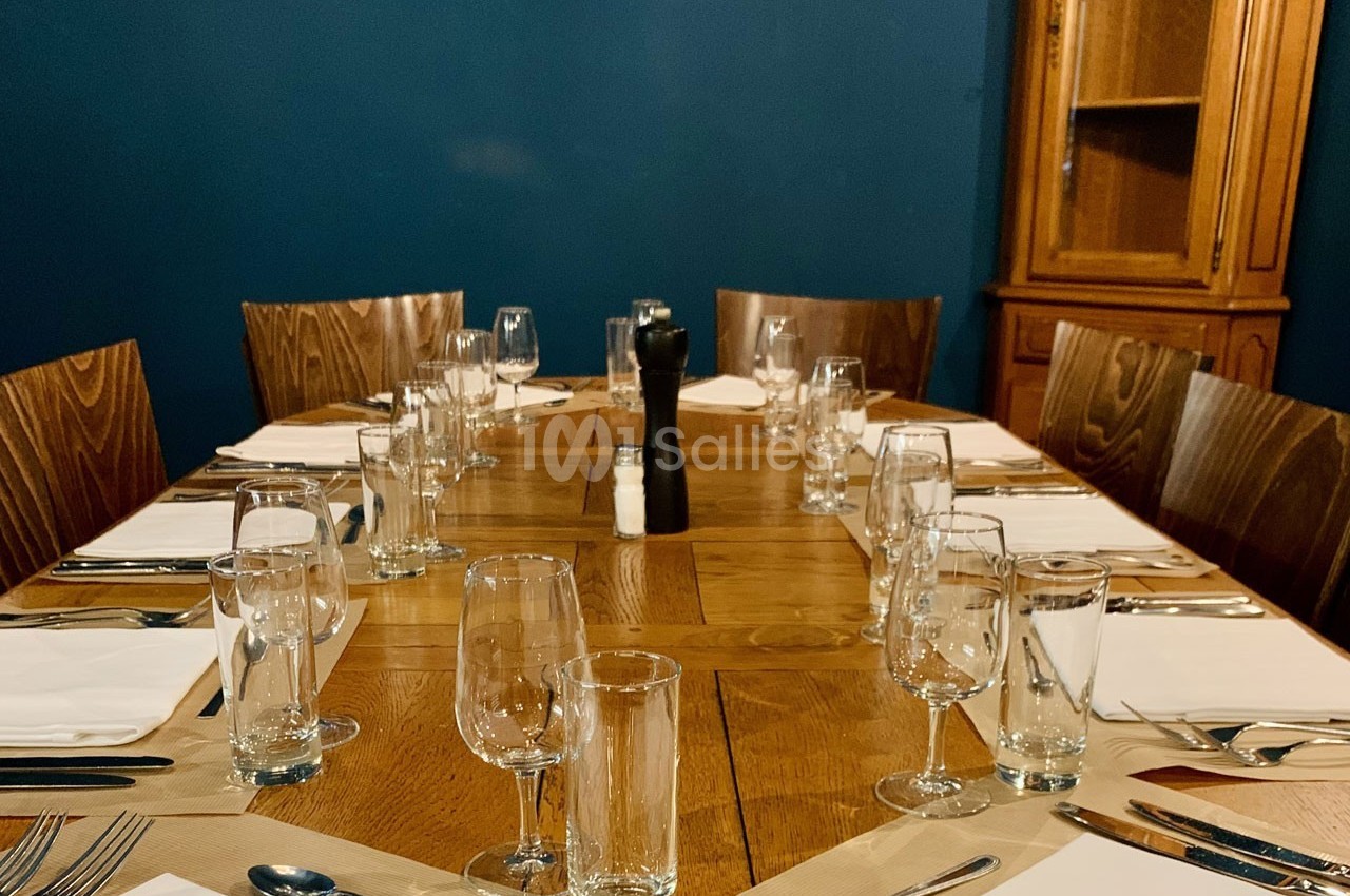Table en bois dressée pour un repas, avec verres, assiettes, couverts et serviettes, entourée de chaises en bois.