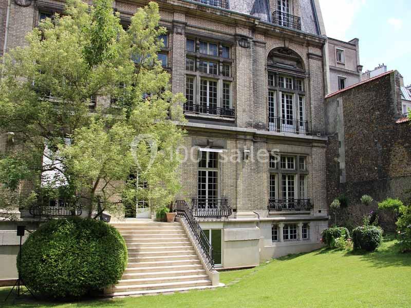 Façade d'un bâtiment en briques avec grandes fenêtres, escalier menant à un jardin verdoyant.