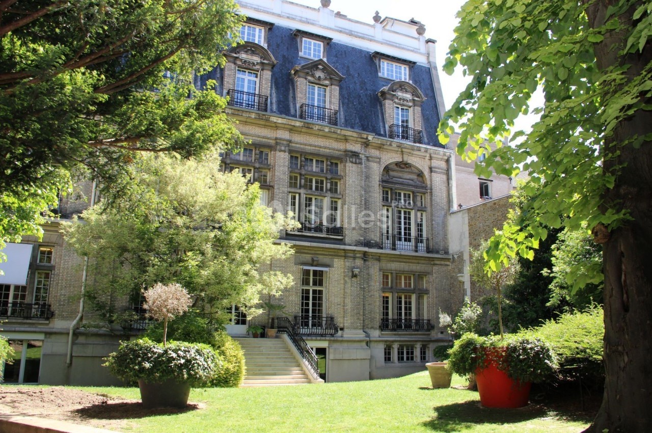 Façade d'un bâtiment ancien entouré d'un jardin arboré avec des pots de fleurs colorés au premier plan.