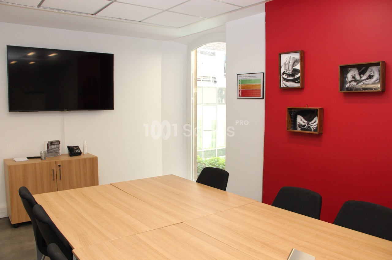 Salle de réunion avec table en bois, chaises noires, mur rouge décoré et écran mural éteint.