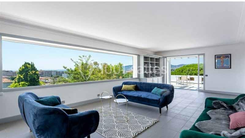 Salon lumineux avec grandes baies vitrées, mobilier moderne, vue dégagée sur des arbres et une terrasse extérieure.