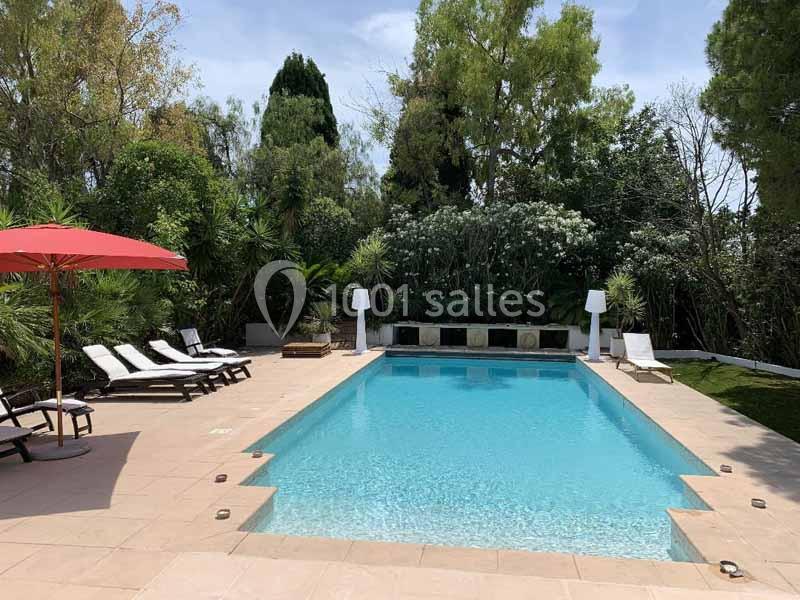 Piscine extérieure entourée de chaises longues, parasol rouge et végétation dense en arrière-plan.