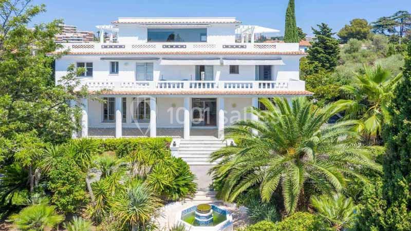 Grande villa blanche entourée de palmiers et d'un jardin verdoyant, avec une fontaine au premier plan.