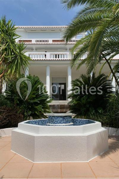 Fontaine octogonale entourée de végétation devant une maison blanche à colonnes sous un ciel bleu.