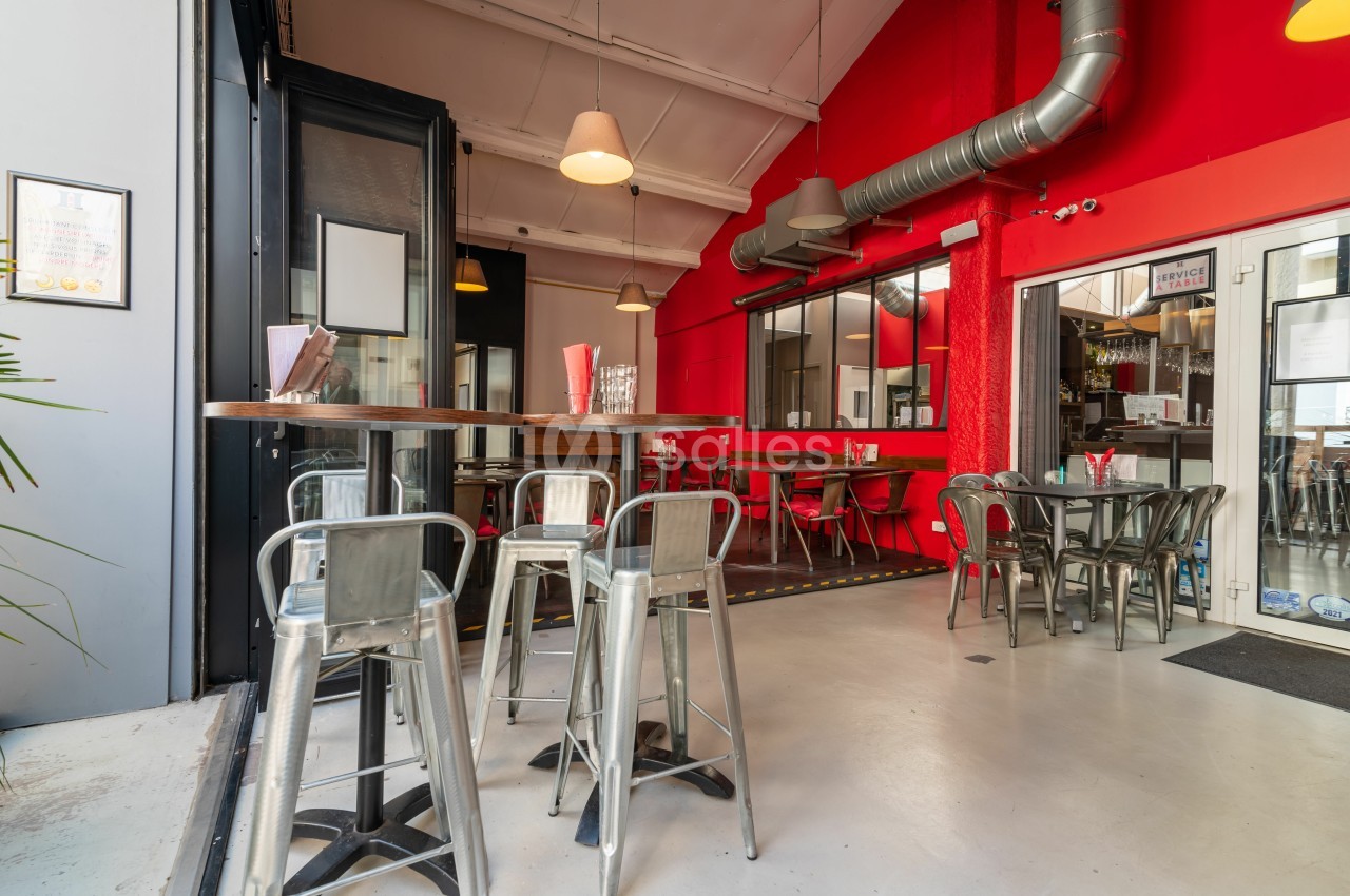 Salle de restaurant moderne avec murs rouges, tables hautes en bois, chaises métalliques et grande baie vitrée.