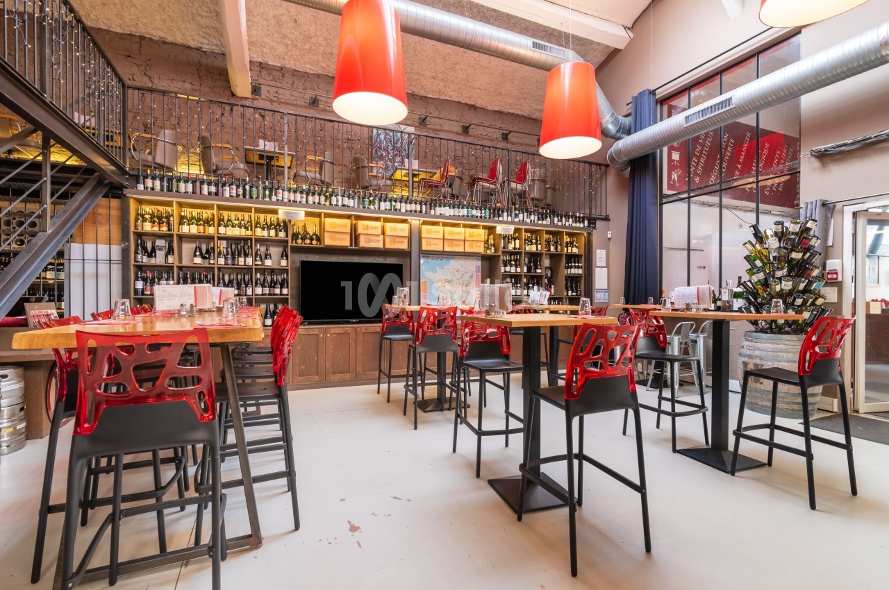 Salle lumineuse avec tables hautes, chaises rouges, étagères remplies de bouteilles de vin et décor industriel.