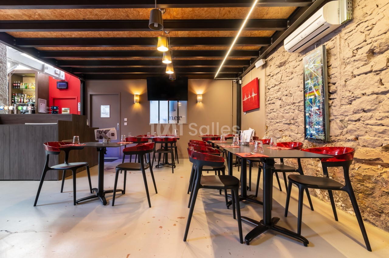 Salle de restaurant moderne avec murs en pierre, tables dressées, chaises rouges et noires, et un bar en arrière-plan.