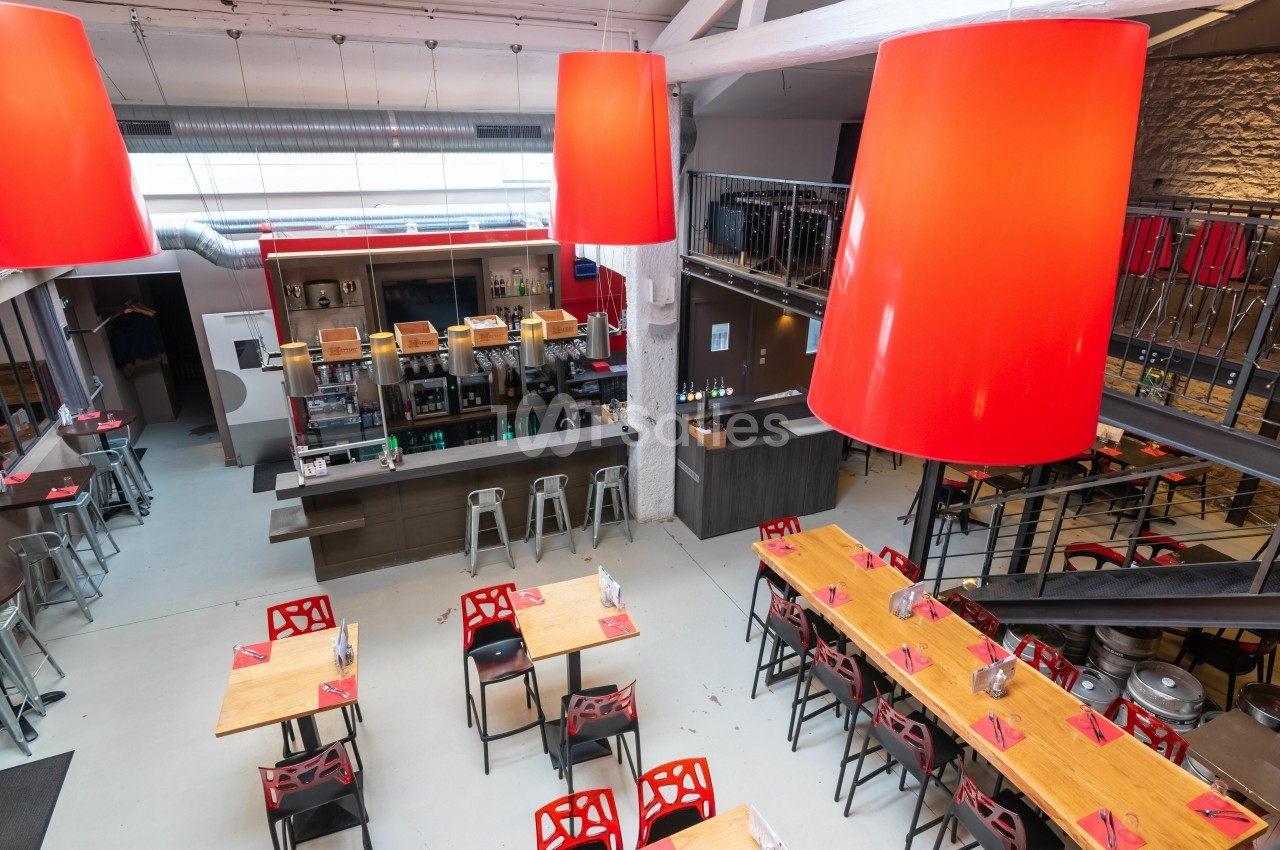 Salle de restaurant moderne avec tables en bois, chaises rouges, bar central et grandes suspensions rouges.