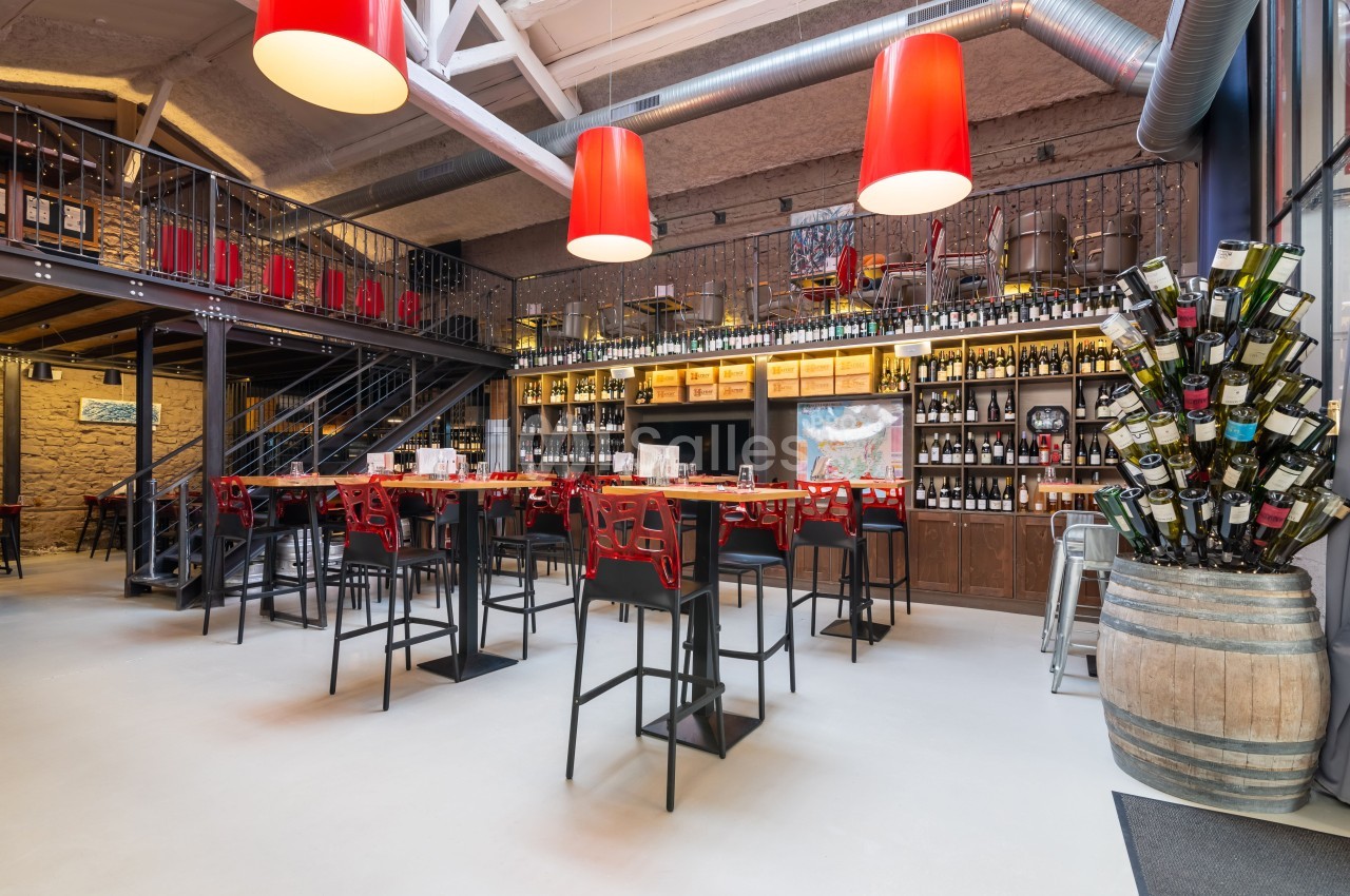 Intérieur d'un bar moderne avec tables hautes, étagères remplies de bouteilles de vin et lampes rouges suspendues.
