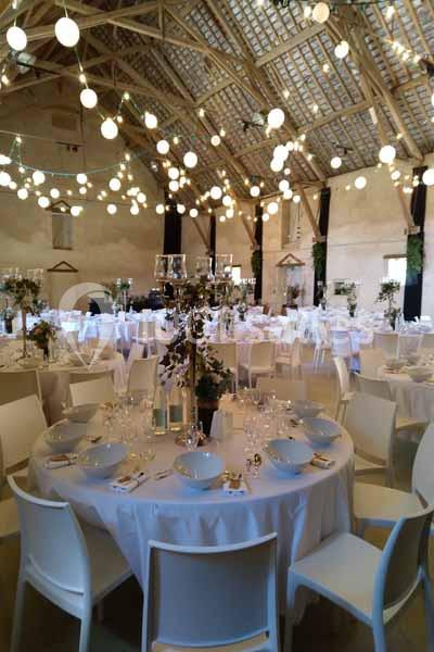 Salle de réception décorée avec des tables rondes dressées, des chaises blanches et des guirlandes lumineuses suspendues.