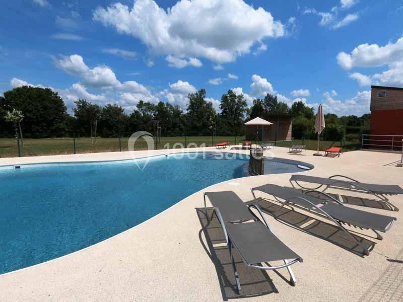 Piscine extérieure avec transats sur une terrasse ensoleillée, entourée d'arbres et d'un ciel partiellement nuageux.