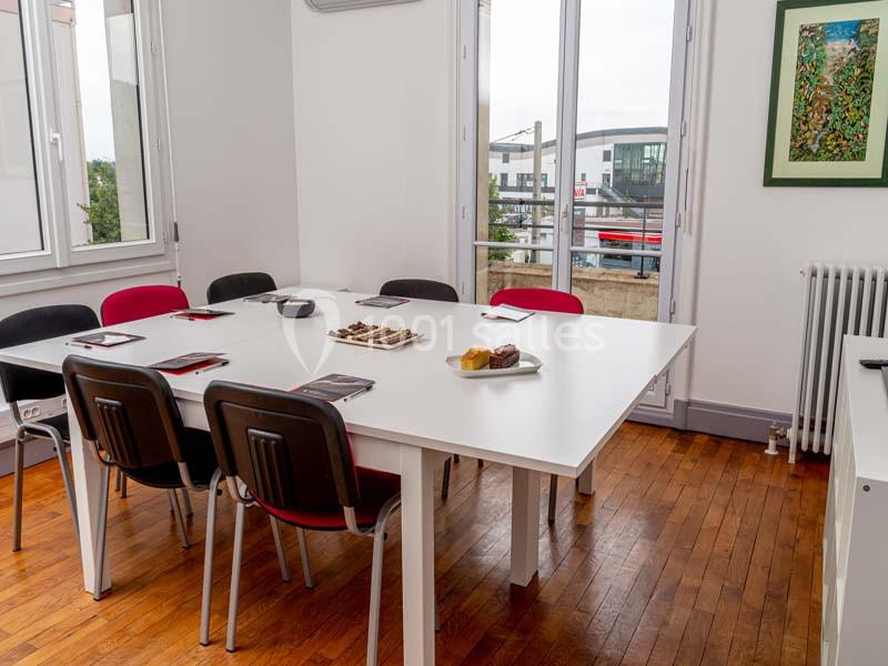 Salle de réunion lumineuse avec table blanche, chaises colorées, documents, fenêtres ouvertes et vue sur l'extérieur.