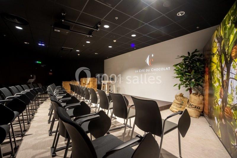 Salle de conférence avec rangées de chaises noires, écran blanc, sacs en toile et plantes en arrière-plan.