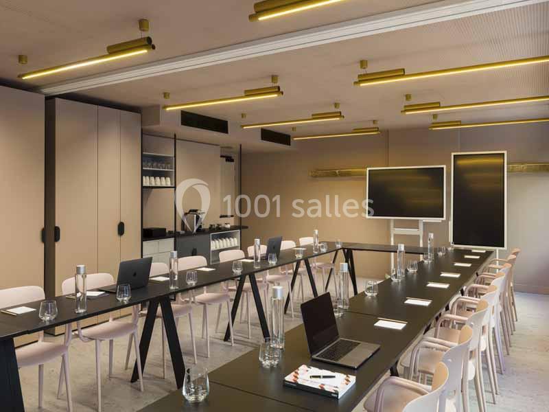 Salle de réunion moderne avec tables noires en U, chaises roses, écran, et matériel de présentation.