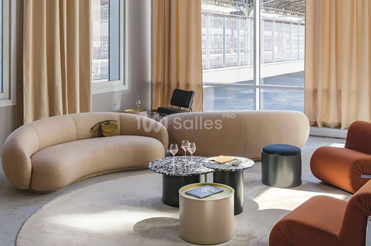 Salon moderne avec canapés beiges, fauteuils orange, tables basses rondes et grandes fenêtres laissant entrer la lumière.