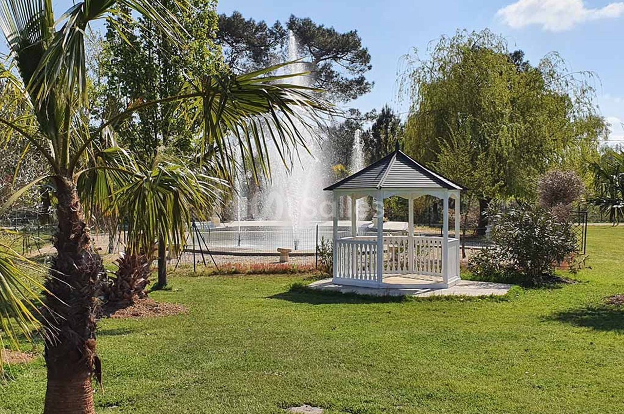 Kiosque blanc dans un parc verdoyant avec une fontaine en arrière-plan sous un ciel ensoleillé.