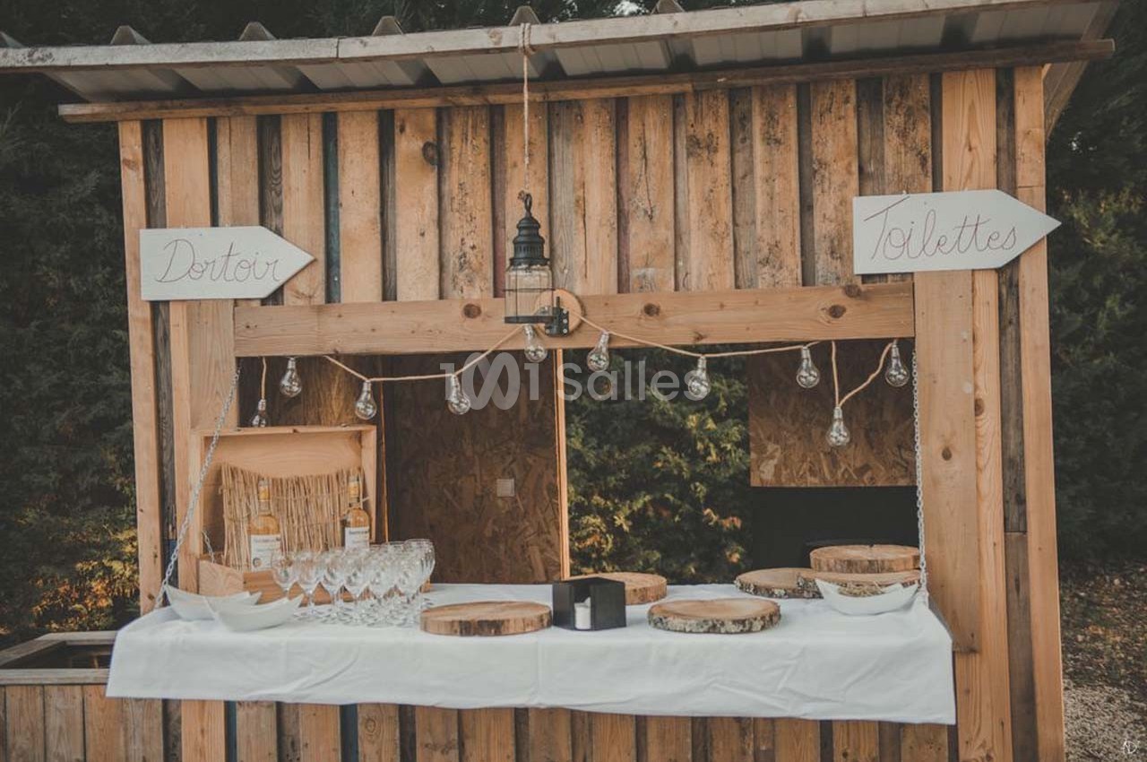 Stand en bois décoré avec des guirlandes lumineuses, proposant des verres et des bouteilles, entouré de panneaux…