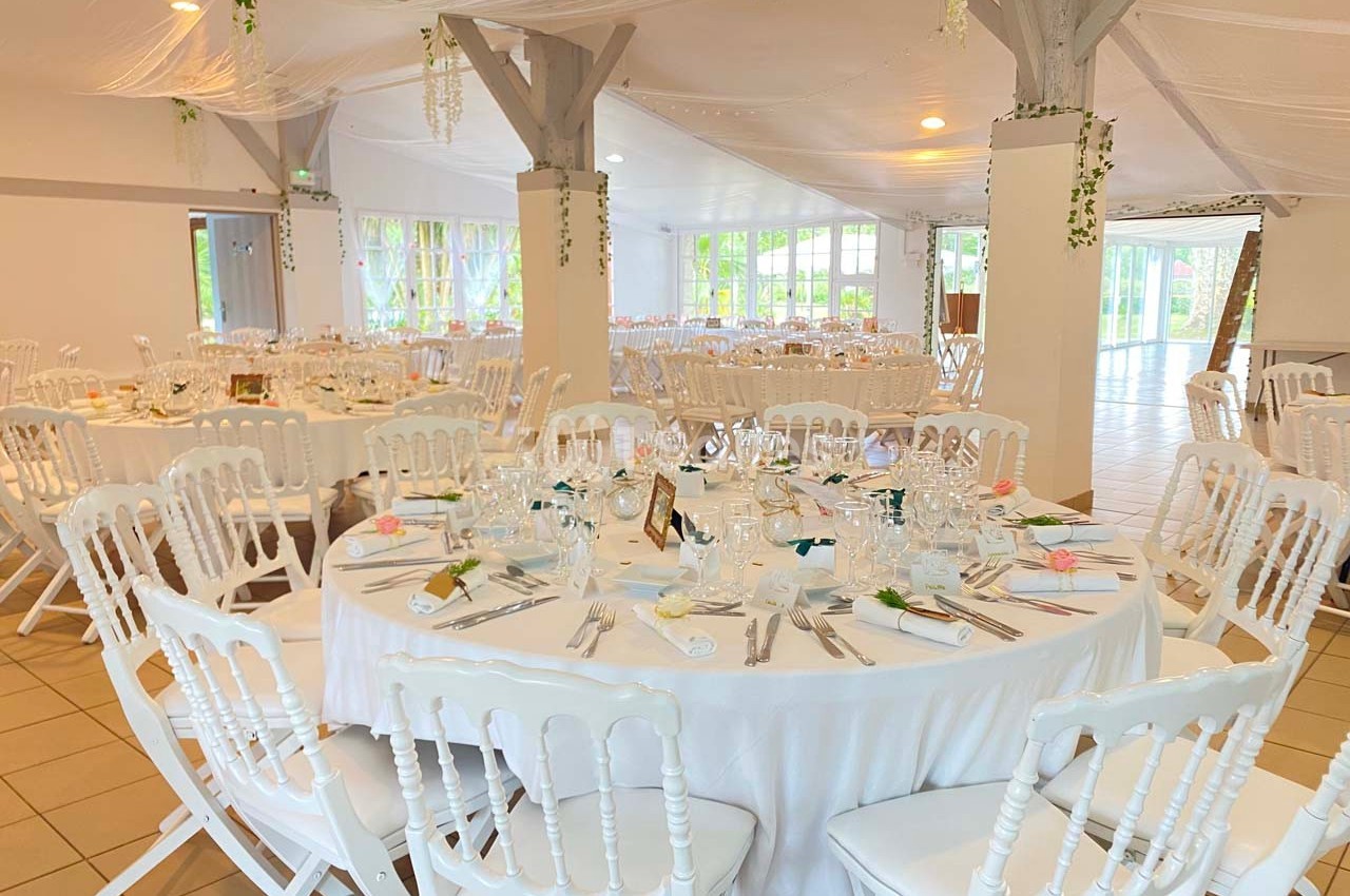 Salle de réception décorée avec des tables rondes dressées, nappes blanches et chaises blanches, ambiance lumineuse.
