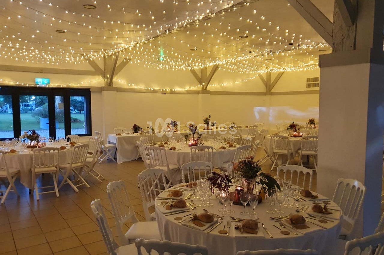 Salle de réception décorée pour un mariage, avec tables rondes dressées et guirlandes lumineuses suspendues au plafond.