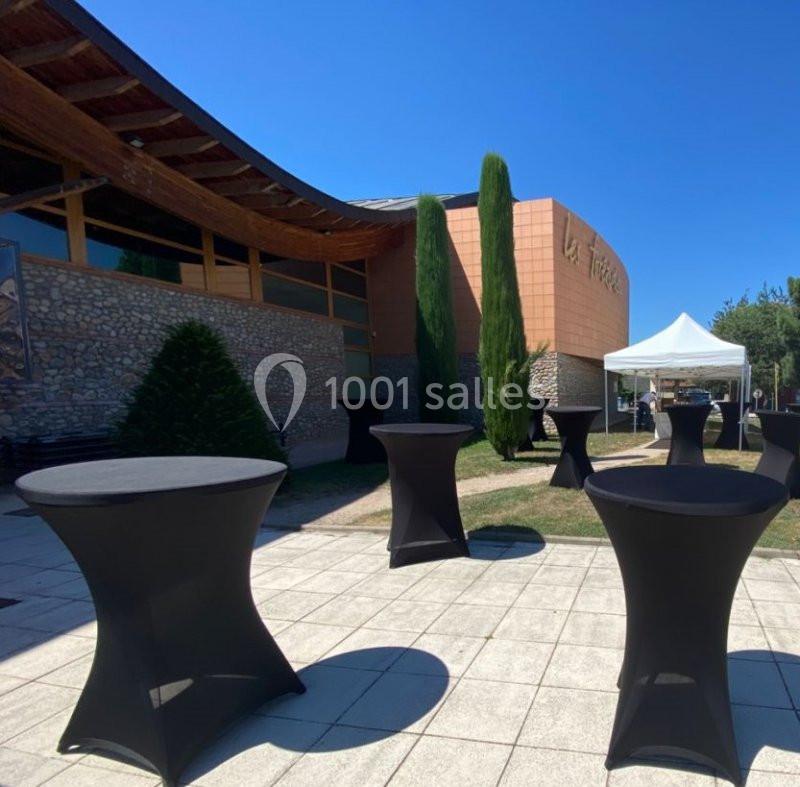 Tables hautes noires disposées en extérieur devant un bâtiment moderne avec une tente blanche et des arbres.