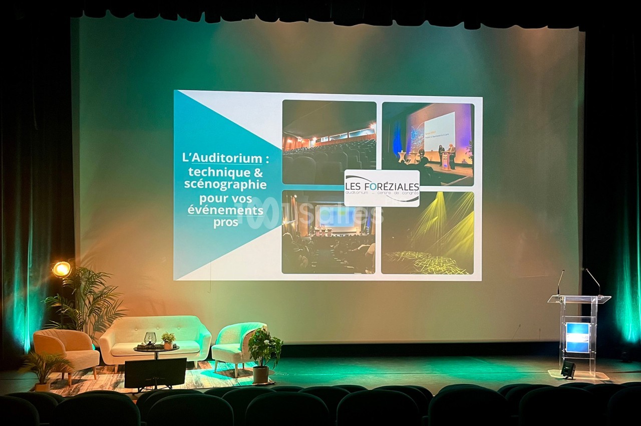 Scène d'auditorium avec fauteuils, écran projetant une présentation et pupitre éclairé pour un événement professionnel.