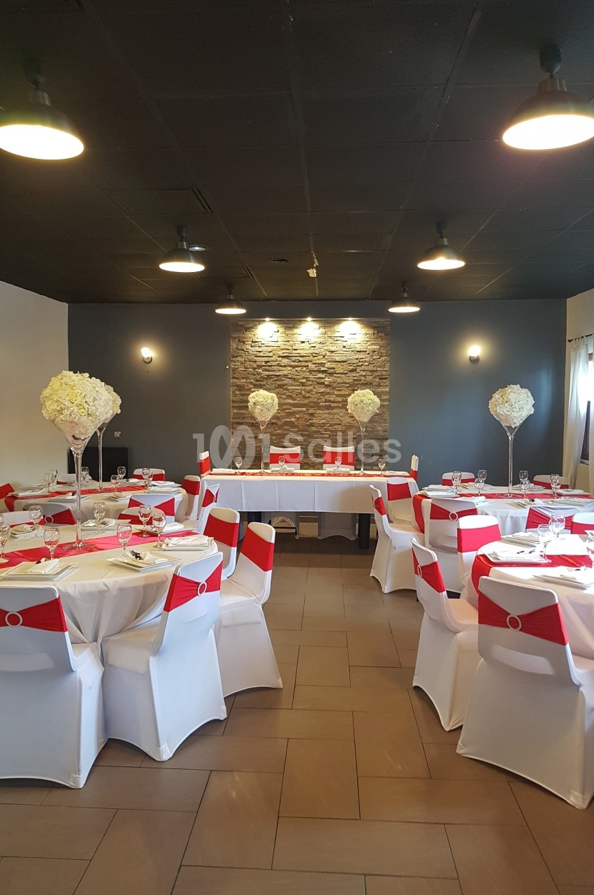 Salle de réception décorée avec des tables rondes, nappes blanches, chaises ornées de rubans rouges et centres de table…