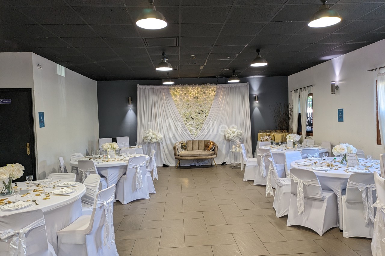 Salle de réception décorée en blanc avec des tables rondes, des chaises habillées et un canapé devant un mur fleuri.