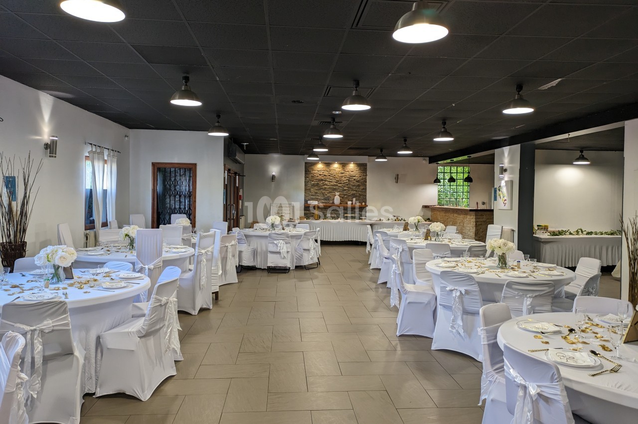 Salle de réception décorée avec des tables rondes dressées, nappes blanches et chaises couvertes, éclairage suspendu.