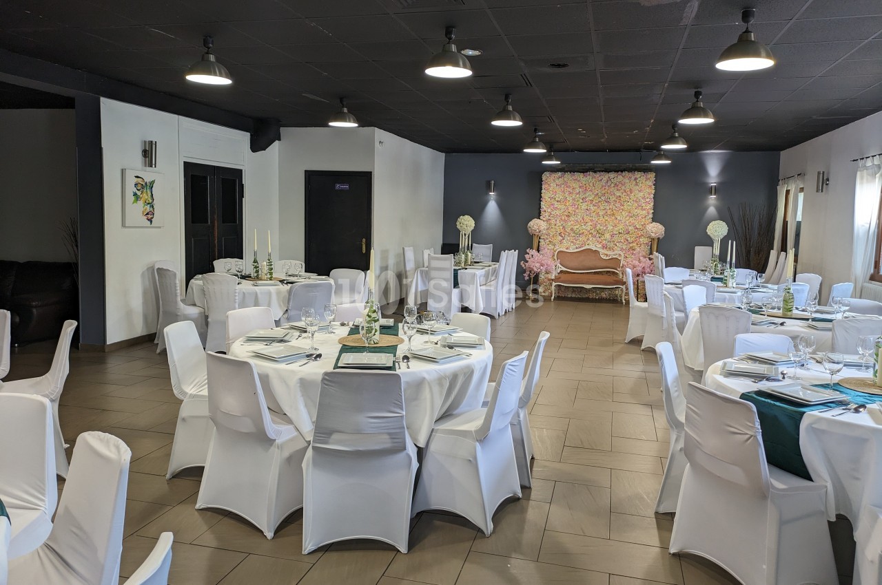Salle de réception décorée avec des tables rondes dressées, chaises blanches et un fond floral coloré.