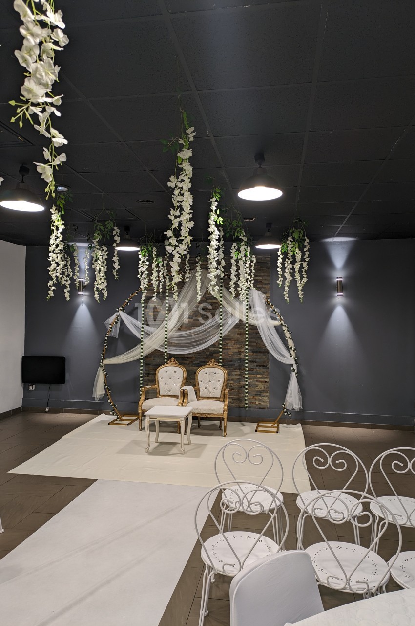 Salle décorée pour un événement avec deux chaises blanches devant une arche ornée de fleurs suspendues et de voilages.