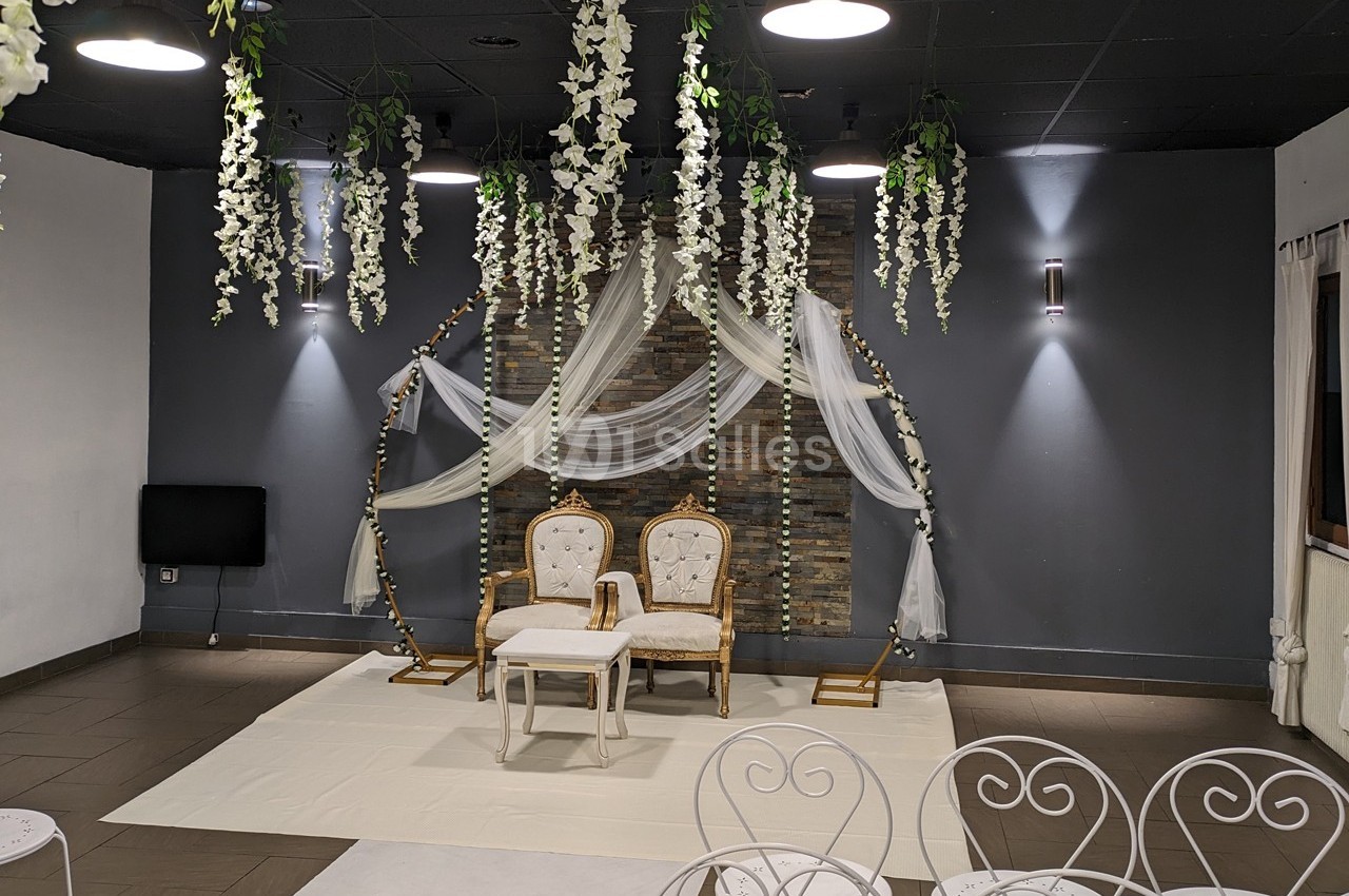 Deux chaises dorées avec une table blanche devant un décor floral suspendu dans une salle sombre.