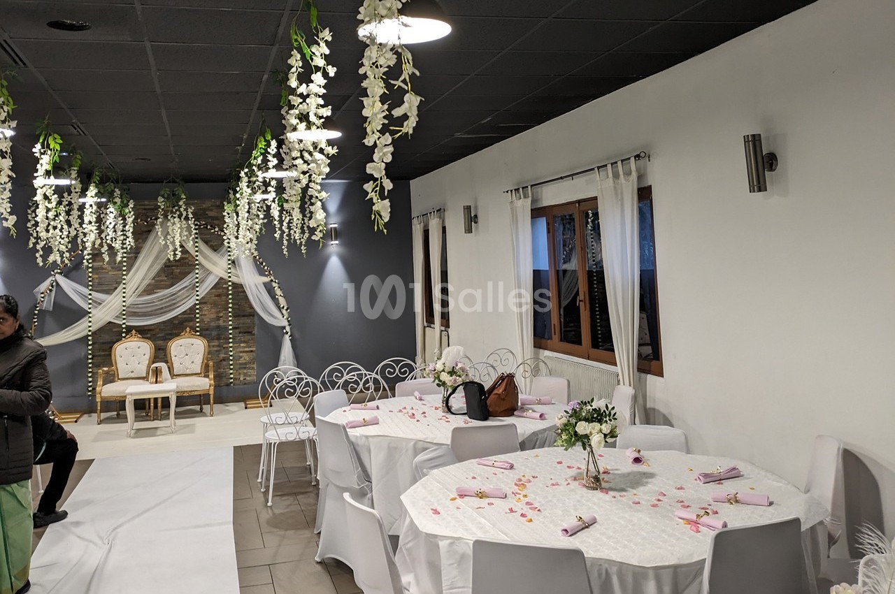 Salle décorée pour un événement avec tables rondes dressées, chaises blanches, fleurs suspendues et espace scénique en…