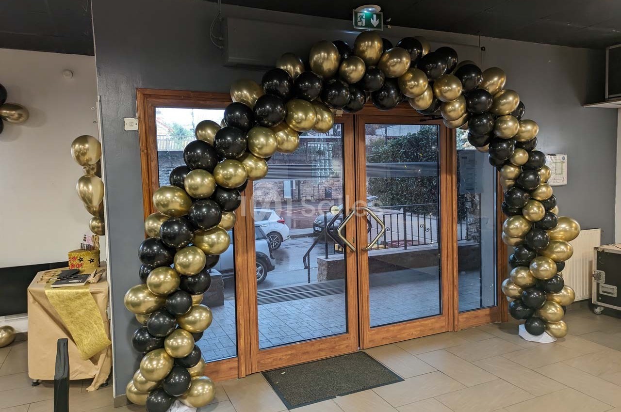 Arche décorative en ballons noirs et dorés installée devant une porte vitrée dans un espace intérieur.