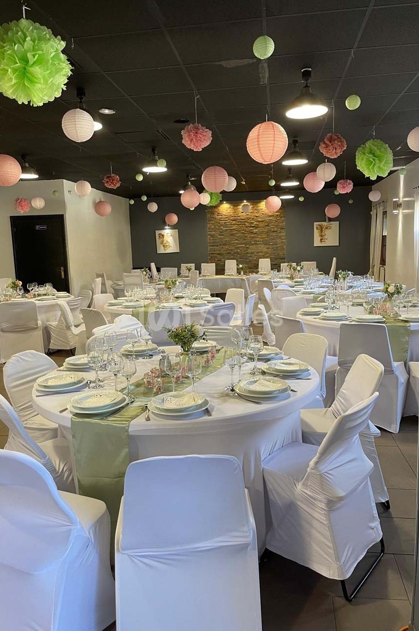 Salle décorée pour un événement, avec tables rondes dressées, nappes blanches et lanternes suspendues au plafond.